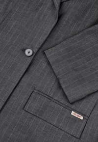 Harmaa raidoitettu pikkutakki, jossa on yksi musta nappi, viistetyt läpät ja pieni etiketti etutaskussa. Pintakuvioitu kangas.