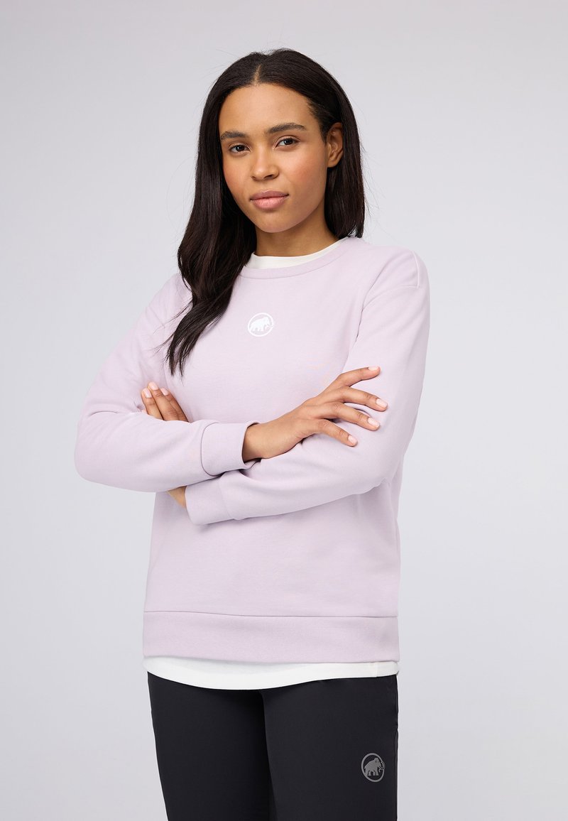Lichtpaarse sweatshirt met een ronde halslijn, geribde manchetten en tailleband; heeft een klein wit logo op de borst. Gedragen over zwarte broek.