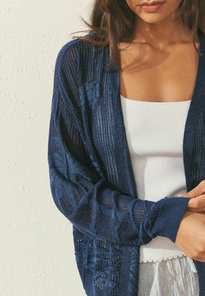 Woman wearing a navy blue knitted cardigan over a white ribbed tank top, with loose hair, standing against a textured white wall.