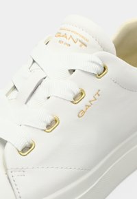 Valget sneakerid valge nahaga, kuldsete silmadega, tekstureeritud valged paelad ja "GANT" bränding kuldse värviga küljepeal. Sile viimistlus ja minimaalne disain.
