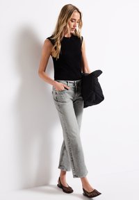 Top sin mangas de canalé negro, vaqueros de denim gris claro con adornos y chaqueta acolchada negra. Modelo lleva zapatos planos de rejilla.