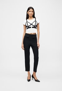 Γυναίκα που φοράει λευκό κοντό τοπ με μαύρη λεπτομέρεια ιμάντα, μαύρο ψηλόμεσο στενό παντελόνι και μαύρες μυτερές ψηλοτάκουνες γόβες, όρθια.