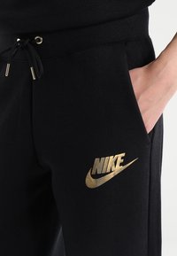 Svarta bomullsjoggers med en guldfärgad Nike-logga på vänster sida, justerbart midjeband och sidofickor. Mjuk, slät textur.