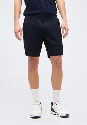 Άνδρας που φοράει σκούρο μπλε σορτς με σχέδιο μέχρι το γόνατο, άσπρες κάλτσες μέχρι τον αστράγαλο και λευκά με μπλε παπούτσια, πάνω σε απλό φόντο.