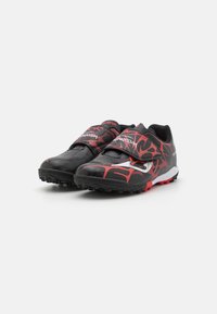 Zapatillas deportivas negras con patrones abstractos rojos, que cuentan con una correa de velcro y una suela de tracción diseñada para un rendimiento óptimo en superficies de césped.