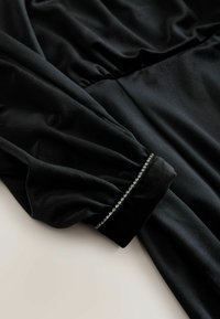 Svart klänning med långa ärmar i mjukt tyg. Ärmsluten pryds av en rad små dekorativa pärlor som ger detalj till den eleganta designen.