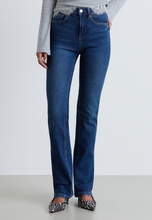 Personne portant un jean bleu droit taille haute, un haut gris à manches longues avec bordure en dentelle, et des chaussures pointues imprimé zèbre noir et blanc.