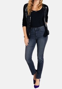Cardigan nero con dettagli in paillettes sopra una canotta nera, abbinato a jeans grigio scuro e tacchi viola, con un design aderente.