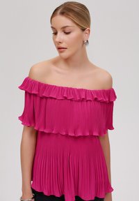 Rosa off-shoulder topp med to volanger, med en plissert tekstur og en flytende silhuett.