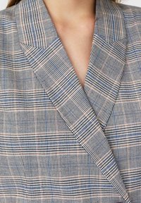 Blazer de cuadros en tonos gris, azul y marrón, con cuello con muesca, tejido texturizado y un ajuste entallado con paneles delanteros superpuestos.