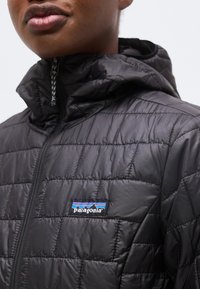 Κοντινό πλάνο ενός ατόμου που φοράει μαύρο καπιτονέ μπουφάν Patagonia με κουκούλα, εμφανίζοντας το μπροστινό φερμουάρ και την ετικέτα με το λογότυπο στο στήθος.