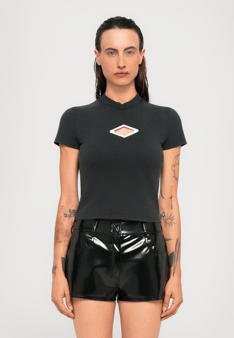 Persona con capelli scuri e bagnati indossa una maglietta nera a maniche corte con un taglio a diamante e pantaloni corti neri lucidi a vita alta, mostrando tatuaggi sulle braccia.