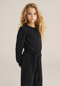 Fekete, hosszú ujjú overall, texturált szövettel, ezüst pöttyökkel, kerek nyakkivágással, és megkötött derékkal az állítható illeszkedés érdekében.