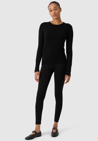 Černý svetr s dlouhými rukávy a kulatým výstřihem, doplněný černými legínami. Hladká textura, přiléhavý střih a rozparky na manžetách.