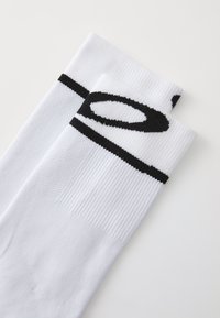 Białe skarpetki o żebrowanej teksturze, z czarną poziomą stripe i logo blisko góry. Materiał wydaje się syntetyczny, zaprojektowany do użytku sportowego.