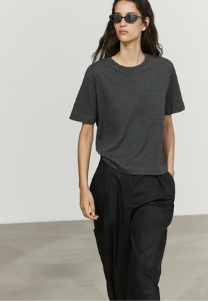 Vrouw draagt donkergrijs T-shirt met korte mouwen, zwarte broek en zwarte zonnebril, loopt binnenshuis tegen een eenvoudige lichte muur.