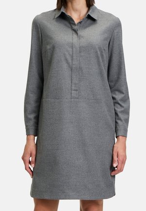 Femme portant une robe-chemise grise à manches longues, longueur genou, avec un col et une patte de boutonnage cachée, debout les bras détendus le long du corps.