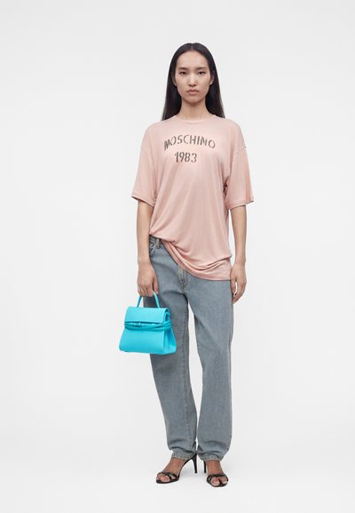 Γυναίκα με ροζ μπλούζα Moschino 1983, φαρδιά ανοιχτό μπλε τζιν, μαύρα σανδάλια με τακούνι, κρατώντας μια μικρή φωτεινή μπλε τσάντα.