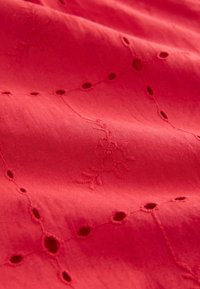 Tessuto rosso con ricami floreali e dettagli a intaglio. La texture appare liscia con motivi di cucitura in rilievo in tutto il tessuto.