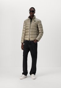 Lys grønn pufferjakke med horisontal quiltet design, glidelås-lukking og stående krage. Kombinert med svarte jeans og hvite joggesko.