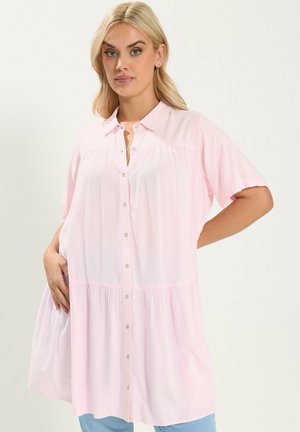 Lyshåret kvinde iført løs, lyserød skjortekjole med korte ærmer, poserer med den ene hånd på hoften og den anden i lommen.