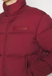 Rode gewatteerde jas met hoge kraag, ritsluiting en reliëf "HILFIGER" logo. Bevat een gestructureerd oppervlak en zijzakken.