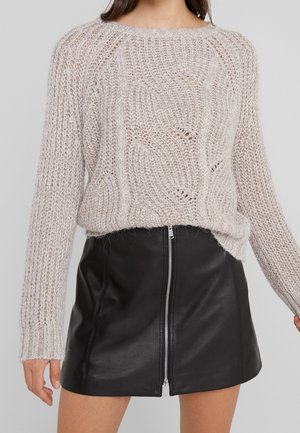 Femme portant un pull en maille épaisse beige et une mini-jupe en cuir noire avec une fermeture éclair à l'avant, debout devant un fond uni.