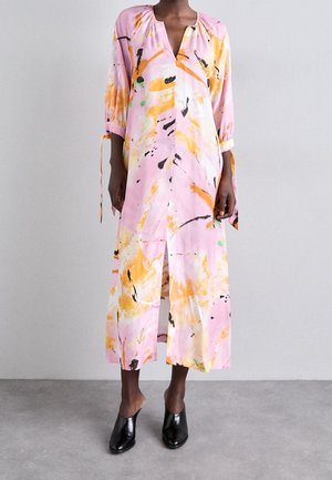 Femme portant une longue robe rose ample avec des motifs abstraits orange, noir et vert, associée à des chaussures noires à enfiler sur un fond uni.
