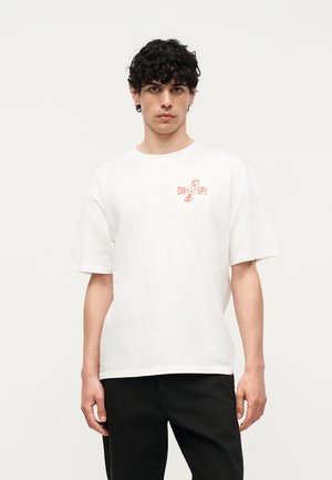 Jeune homme aux cheveux bouclés et foncés portant un t-shirt blanc à manches courtes avec l'inscription orange "EDWIN SUPPLY" et un pantalon noir sur fond uni.