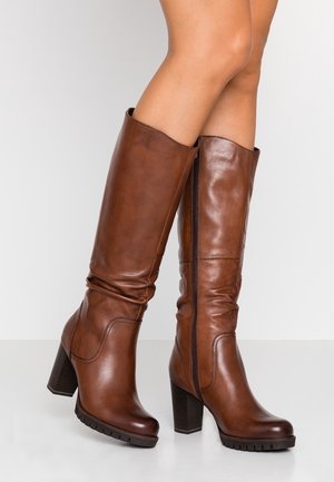 Brązowe skórzane kozaki za kolano na słupku z bocznymi zamkami, noszone na skrzyżowanych nogach na jednolitym tle.