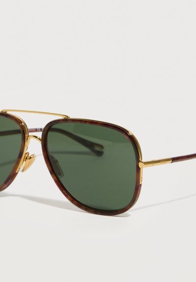 Γυαλιά ηλίου με σκουρόχρωμους πράσινους φακούς, μεταλλικά σημεία σε χρυσό και πλαίσια με μοτίβο χελώνας, εμφανίζονται σε κοντινό πλάνο πάνω σε λευκό φόντο.