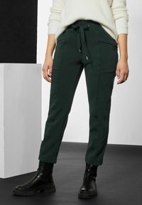 Mörkgröna skräddarsydda byxor med dragsko i midjan, två sidofickor och avsmalnande passform, i kombination med svarta ankelstövlar.