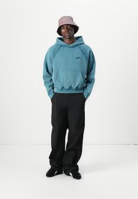 Blauer, übergroßer Hoodie mit einer Vorderansicht, kombiniert mit schwarzen weit geschnittenen Hosen und einem hellrosa Bucket-Hut. Schwarze Schuhe runden das Outfit ab.