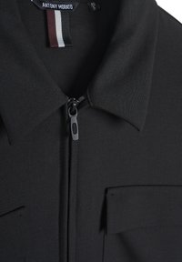 Maglietta nera con zip e colletto a punta, due tasche frontali, tessuto liscio e un'etichetta che mostra il nome del marchio e la taglia.