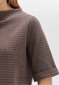 Top marrone con texture e motivo a griglia, maniche corte arrotolate e colletto alto. Il materiale appare morbido e leggermente elasticizzato.
