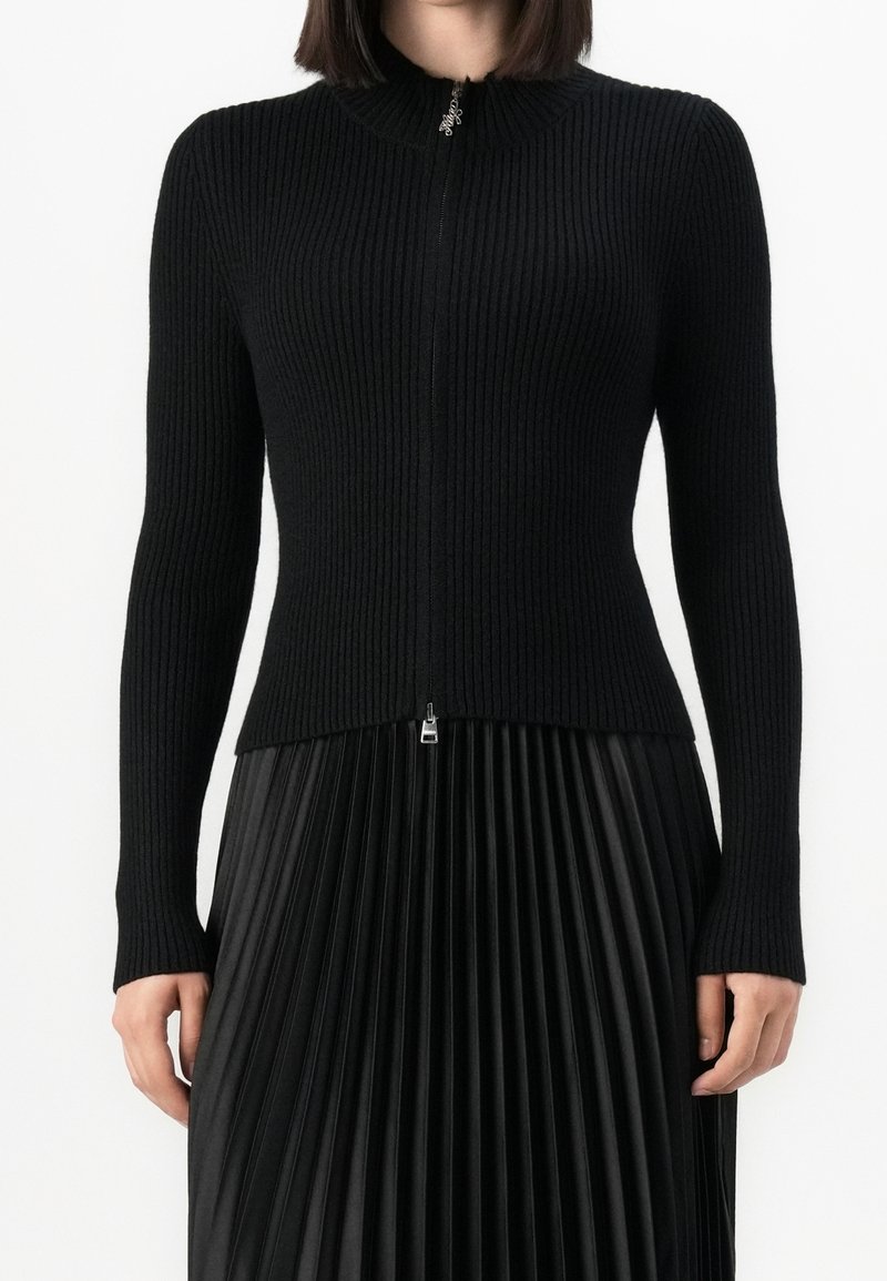 Femme portant un pull noir côtelé ajusté à fermeture éclair, associé à une jupe plissée noire taille haute.