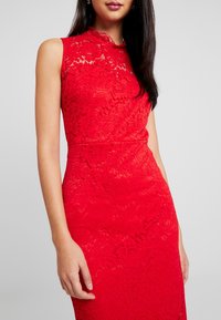 Robe rouge en dentelle avec un col haut festonné, un design sans manches, une silhouette ajustée, et des motifs floraux complexes sur toute la surface.