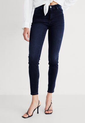 Jean skinny en jean extensible, de couleur denim foncé, avec une taille haute. Présente une finition lisse et un léger délavage au niveau des genoux. Porté avec des talons noirs.
