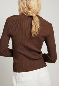 Kvinna med vågigt blont hår som bär en ribbad brun tröja, vänd bort, med en ljus bakgrund och vita byxor synliga.