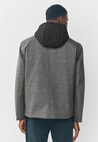 Chaqueta de tela gris con un acabado texturizado, que presenta una capucha acolchada negra. El diseño incluye mangas largas y un dobladillo recto.