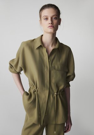 Młoda kobieta o blond włosach nosi luźną oliwkowozieloną koszulę z podwiniętymi rękawami i dopasowane spodnie na jasnym, jednolitym tle.