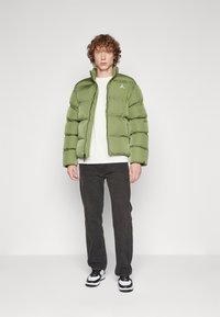 Giacca a vento verde con design a costine orizzontali, zip completa e tasche laterali. Indossata sopra una maglietta bianca, abbinata a jeans scuri e sneakers.