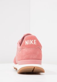 Růžové semišové tenisky s polstrovaným podpatkem, na kterých je bílý logo "NIKE" a světle růžové akcenty na svršku a podešvi; texturovaný povrch.