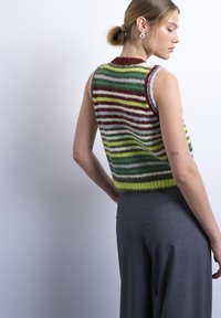 Γυναίκα με ξανθά μαλλιά σε κότσο που φοράει ασημένια σκουλαρίκια, πολύχρωμο ριγέ χνουδωτό αμάνικο πουλόβερ και γκρι παντελόνια, στραμμένη πλαγίως.