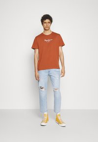 T-maica v rjavi barvi z belim logotipom, svetlo modri raztrgani jeans in rumeni superge, oblečeni s prozornimi očali.