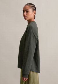 Giovane donna con capelli intrecciati che indossa un top a maniche lunghe verde scuro e una gonna plissettata verde chiaro, in piedi di lato contro uno sfondo semplice.