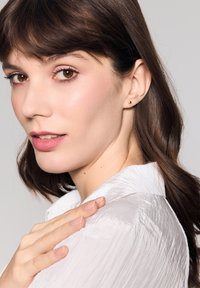 Złota kolczyk wkręcany, przeźroczysta biała koszula z teksturowanym materiałem, gładka skóra z delikatnymi refleksami, brązowe oczy i długie, proste brązowe włosy.