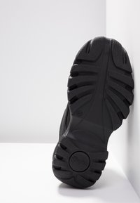Semelle de chaussure noire inclinée contre un angle blanc, présentant des rainures profondes et un profil texturé avec le logo "Buffalo" près du talon.