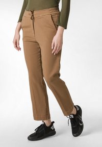 Hnědé tapered chinos s elastickým pasem na tkaničku, bočními kapsami a hladkým textilním vzhledem, doplněné černými sportovními botami.