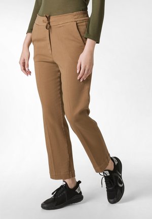 Hnědé tapered chinos s elastickým pasem na tkaničku, bočními kapsami a hladkým textilním vzhledem, doplněné černými sportovními botami.